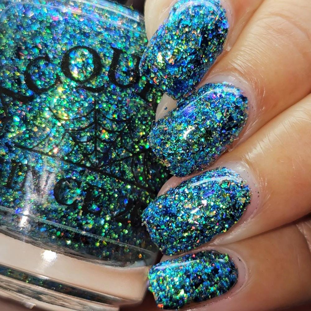 Close-up of a hand with nails painted in a highly reflective blue and green holographic glitter nail polish, next to the bottle of polish.