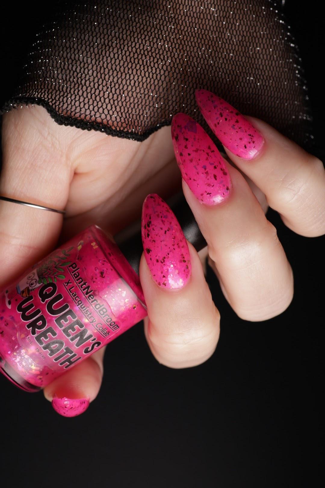 A close-up of a hand with light skin with long, bright pink glittery press-on nails holding a bottle of neon pink nail polish labeled 'Queen's Wreath' PlantNerdBroom against a black background.