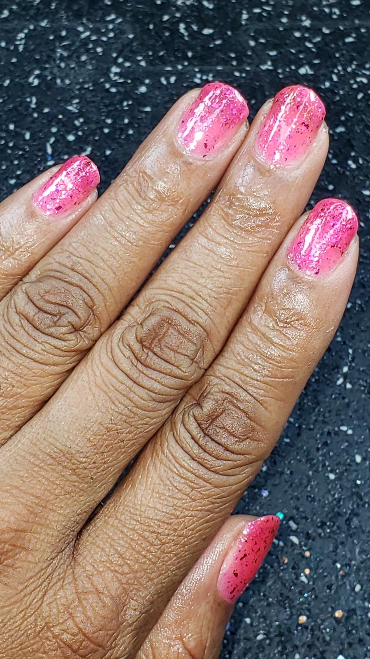 hand with dark skin and short natural nails wearing a sheer pink jelly flake polish against a blue sparkly background