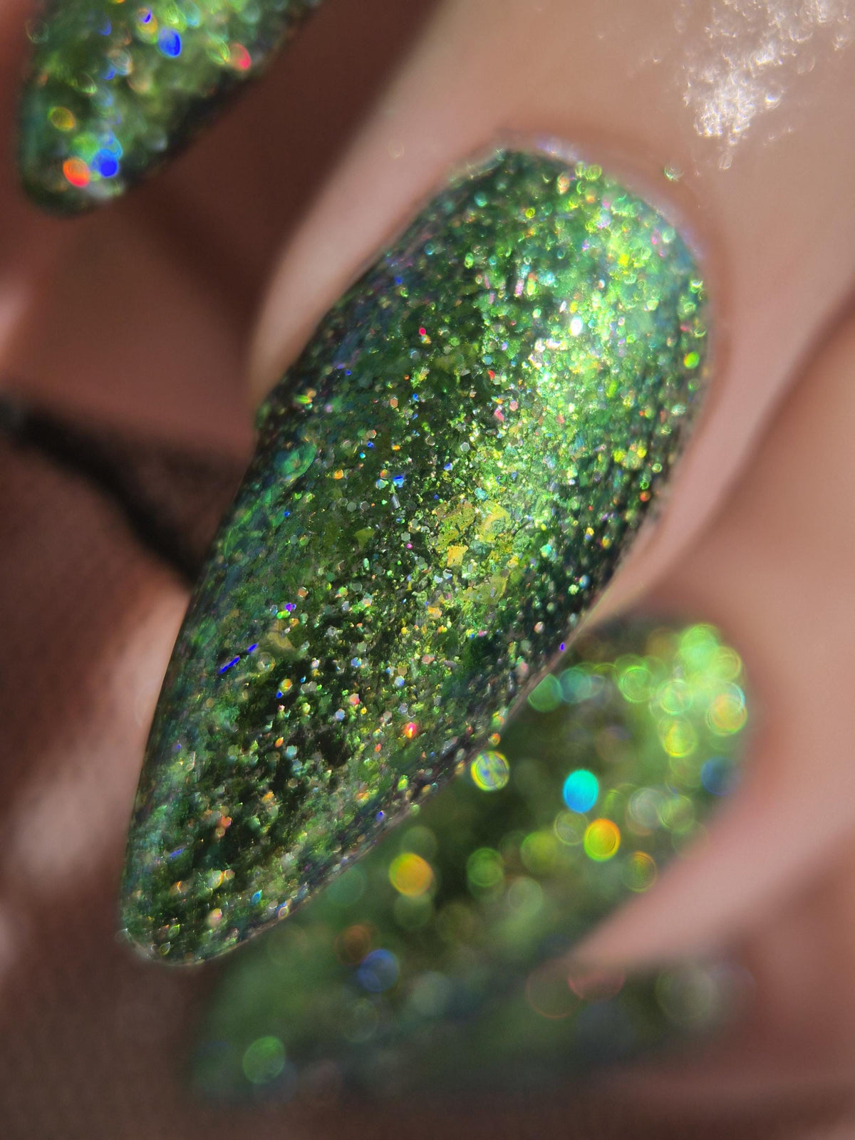 Close-up of a manicured fingernail with long almond-shaped tips covered in dense green glitter and holographic sparkle, shown on a hand.