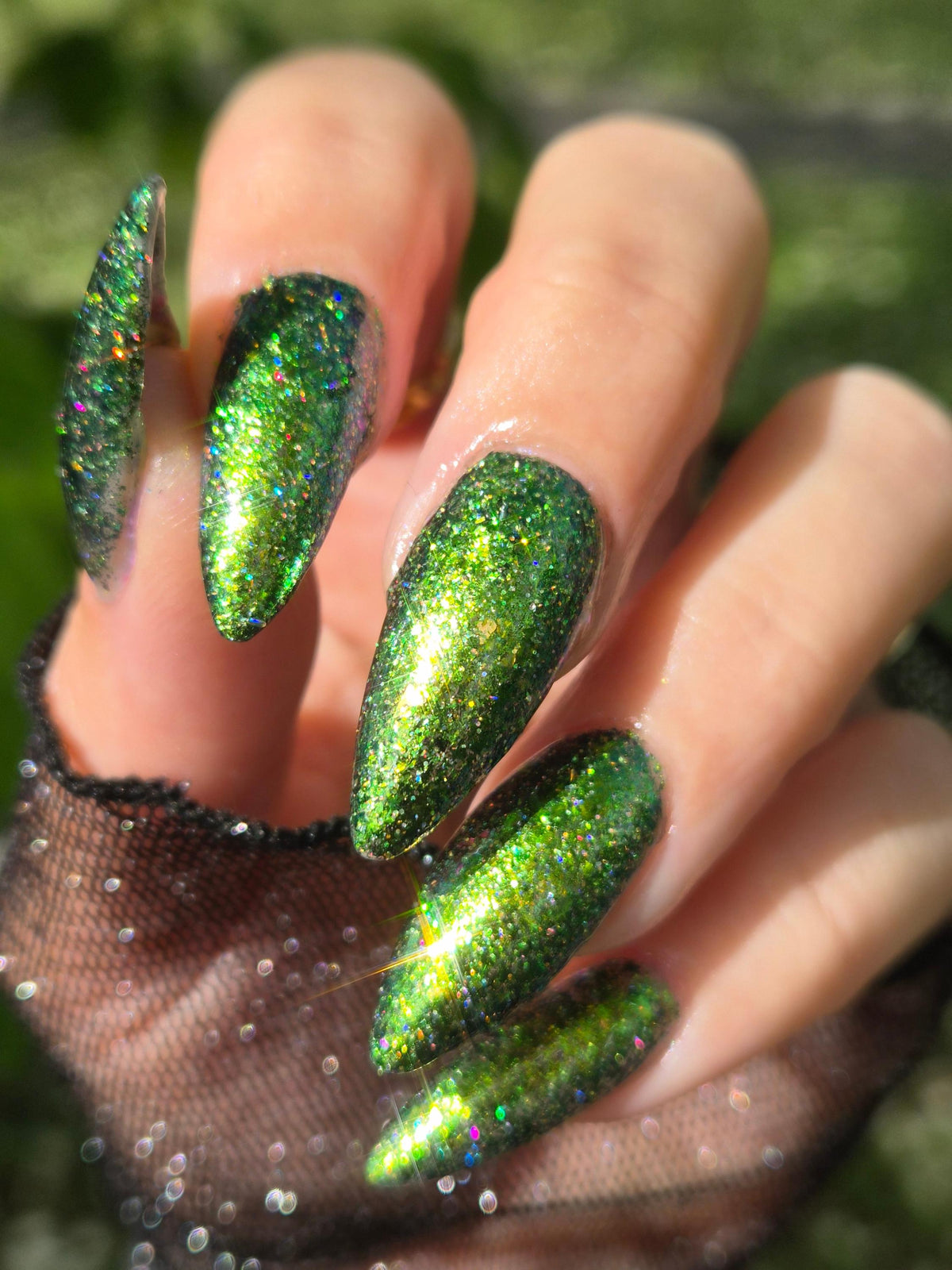 A close-up of a hand with long, almond-shaped nails painted in a sparkling metallic green glitter polish, with a sheer black glove partially visible at the wrist against a blurred green outdoor background.