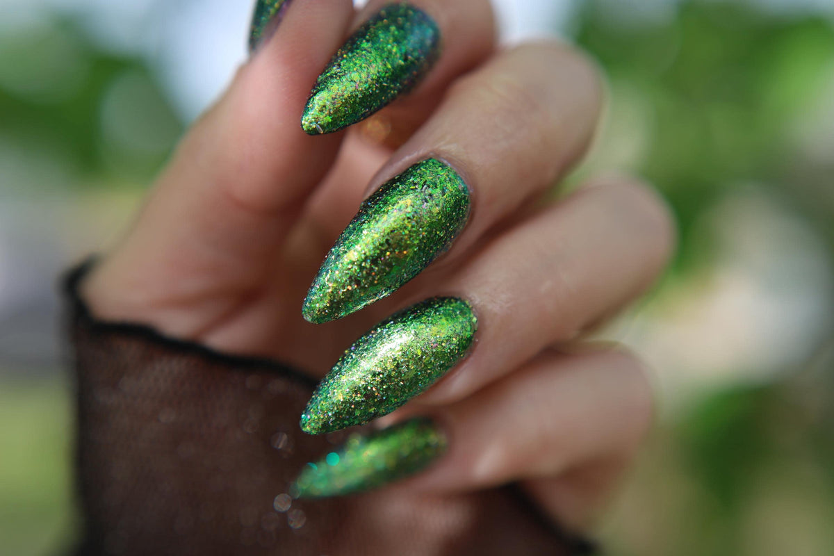 A close-up of a hand with long, almond-shaped nails painted in a sparkling metallic green glitter polish, set against a blurred green outdoor background.