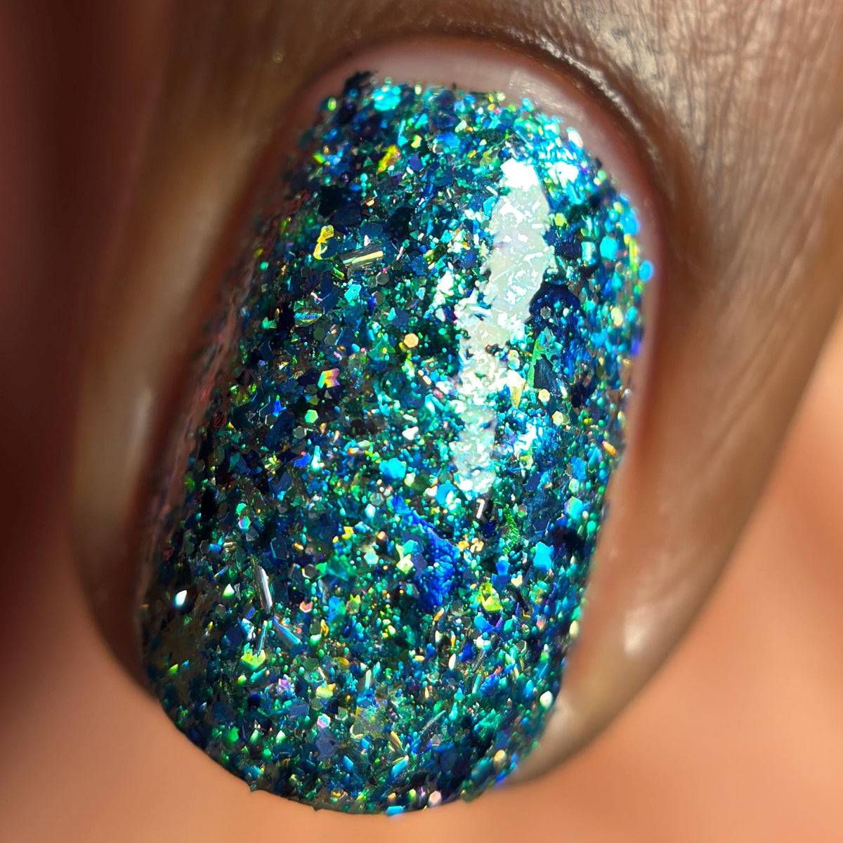 A close-up of a fingernail painted with dense blue and green holographic glitter polish, showing iridescent flecks reflecting light.
