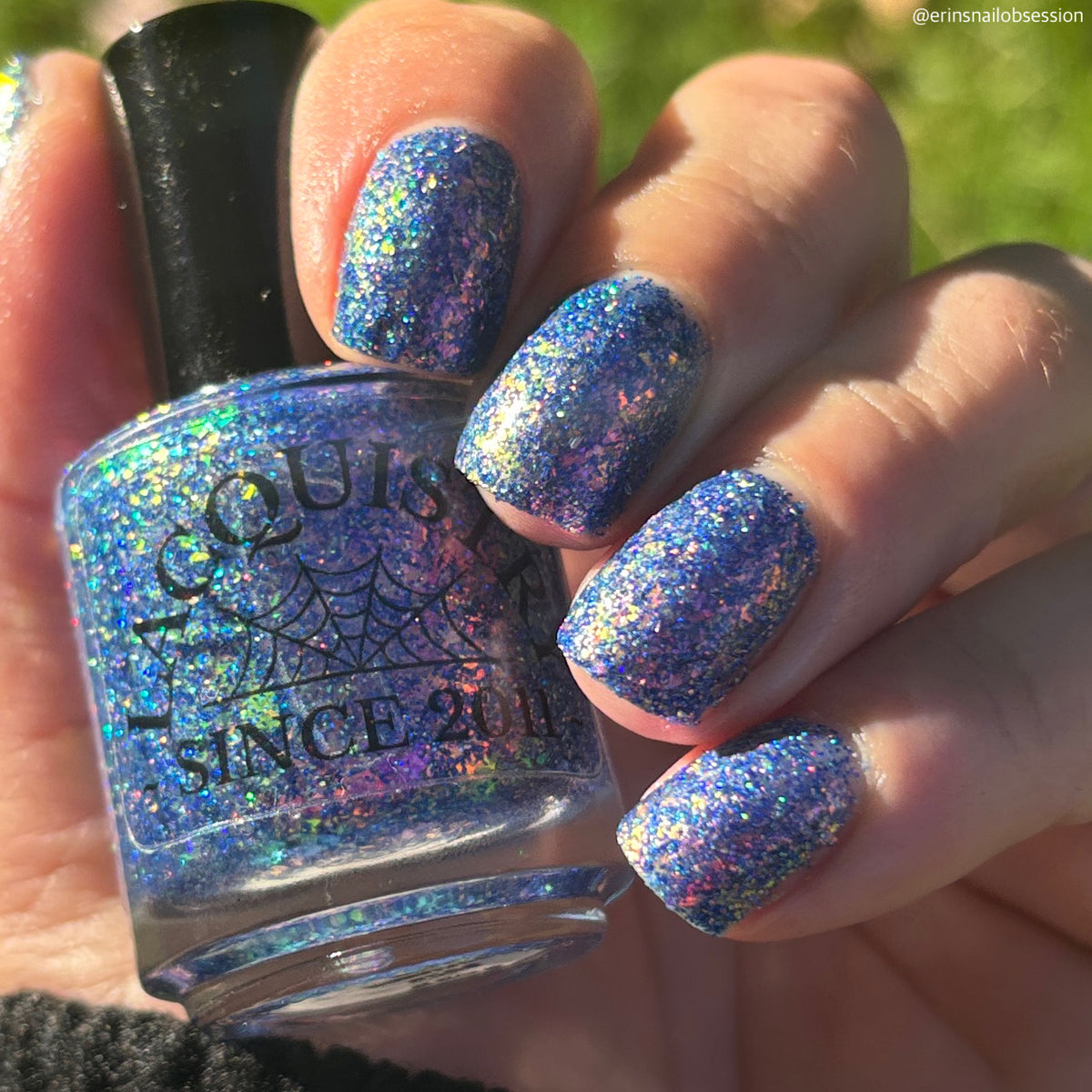 A hand with fingernails painted in a glittery blue nail polish holding a bottle of the same nail polish. The polish has a holographic, iridescent shimmer that reflects purple, green, and orange.