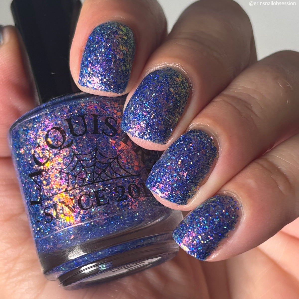 A close-up of a hand holding a bottle of nail polish. The nails are painted with a glittery blue holographic polish that shifts between blue, purple, and gold, matching the color of the polish in the bottle.