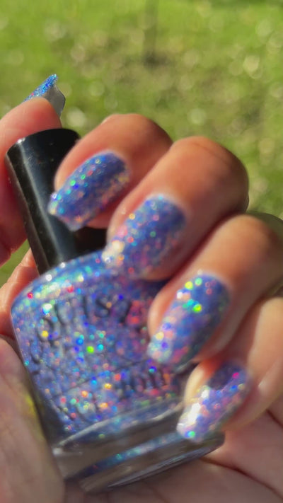 Bright Blue and purple glitter  flakie Lacquistry polish Thriver Not survivor sparkling in the sun with a grassy blurry background
